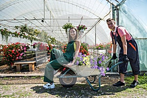 Young happy couple having fun at greenhouse while man rides wife on a wheelbarrow
