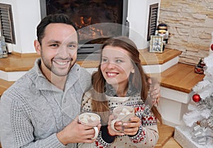 young happy couple with hot cocoa drink in comfy sweaters sitting near fireplace