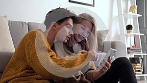 Young happy couple laughing, holding smartphone using mobile cell phone at home.