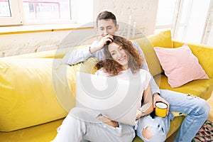 Young happy couple lies on a yellow sofa with a laptop in their apartment, online shopping and internet technology for home