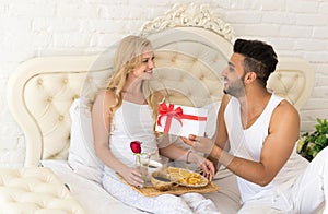 Young Happy Couple Lying In Bed, Hispanic Man Give Woman Surprise Present Envelope With Ribbon, Anniversary Celebration