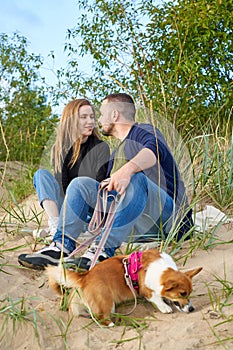 Young happy couple of man and woman with corgi dog sit at sand. Male kissing beautiful female.