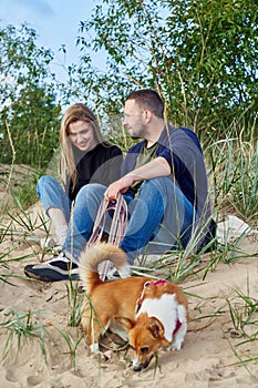 Young happy couple of man and woman with corgi dog sit at sand. Two persons in casual clothes