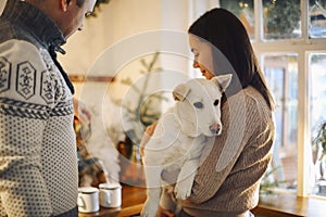 Young happy couple man and woman holding cute white dog and hugging while standing in kitchen decorated for Christmas