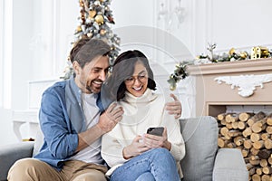 Young happy couple man and woman sitting at home on sofa hugging near Christmas tree and using mobile phone, shopping