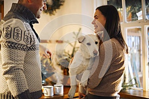 Young happy couple man and woman holding cute white dog and hugging while standing in kitchen decorated for Christmas