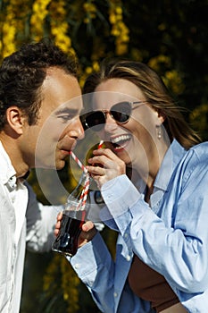 Young happy couple outdoors drinking cold drink from glass bottle with straw. Side view of man and woman drinking soft