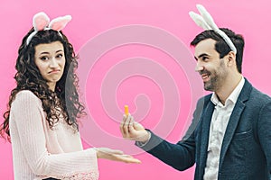 Young happy couple on the pink background. On the head is a rabbit ears. A young man holding a small carrot in his hands