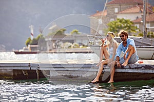 A young happy couple is relaxing with the drink while sitting on the dock on the seaside with their legs in the water. Love,