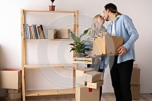 Young happy couple in a room with moving boxes in a new home. Husband and wife kiss standing with boxes