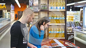 Young happy couple with shopping cart coming up to the the freezer in grocery store and picking packages with frozen