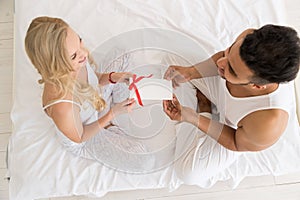 Young Happy Couple Sitting In Bed, Hispanic Man Give Woman Surprise Present Envelope With Ribbon, Anniversary