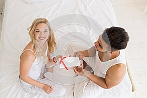 Young Happy Couple Sitting In Bed, Hispanic Man Give Woman Surprise Present Envelope With Ribbon, Anniversary