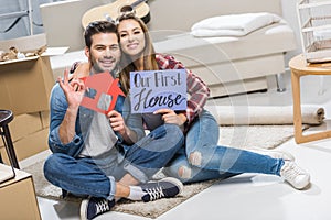 Young happy couple sitting on floor and showing signs with house and our first
