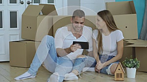 Young happy couple sitting on floor using tablet computer to find transport service for reloction in their new home