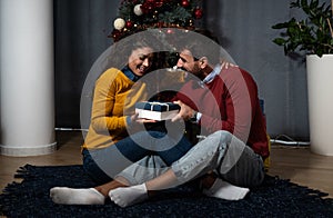 Young happy couple sitting at home in front of the Christmas tree decorated with decorations exchanging gifts for the New Year