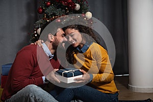 Young happy couple sitting at home in front of the Christmas tree decorated with decorations exchanging gifts for the New Year