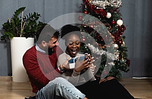 Young happy couple sitting at home in front of the Christmas tree decorated with decorations exchanging gifts for the New Year