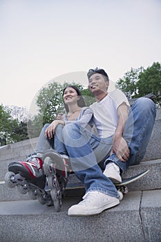 Young happy couple sitting and resting on concrete steps outside with a skateboard and roller blades