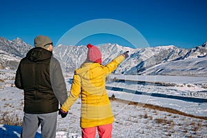 Young happy couple in snowy mountains with copy space. Guy and girl in bright winter clothes look at the snowy peaks. Christmas