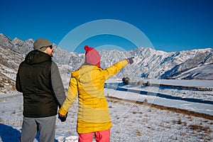 Young happy couple in snowy mountains with copy space. Guy and girl in bright winter clothes look at the snowy peaks. Christmas