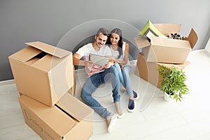 Young happy couple with tablet PC and moving boxes sitting on floor at new home