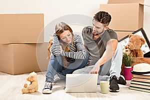 Young happy couple with tablet PC and moving boxes sitting on floor at new home