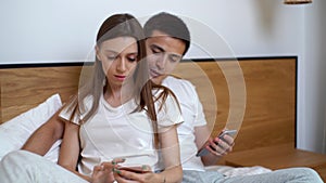 Young happy couple talk laugh looking at smartphone using funny apps sit on bed