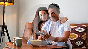 Young happy couple use smart phone together at home sitting on the couch - cheerful girl and boy interracial people using