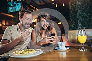 Young happy couple using phone and eating pizza in a restaurant. Selective focus