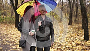 Young happy couple walking together in autumn park holding a colorful umbrella. Attractive woman with red hair is