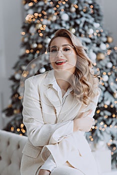 Young happy european businesswoman wearing glasses sitting at workplace with decorated Christmas tree behind, smiling woman