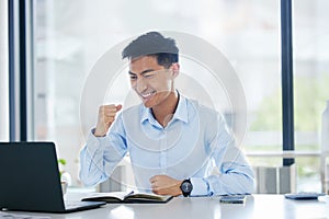 Young happy and excited asian businessman cheering with his fists working on a laptop sitting in an office alone at work