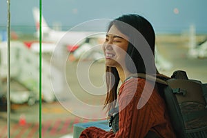 Young happy and excited Asian Korean student woman with backpack at airport departure lounge watching aircraft through glass