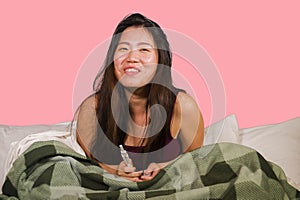 Young happy and excited Asian Korean woman in bed holding pregnancy test checking surprised positive pregnant result smiling ecsta