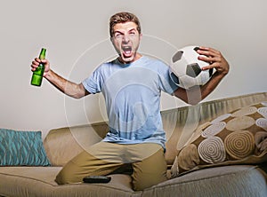 Young happy excited and crazy football fan man holding soccer ball celebrating team scoring goal and victory watching game on tele