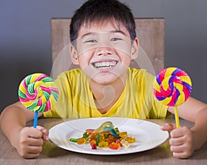 Young happy and excited male kid smiling cheerful eating dish full of candy and lollipop sitting at table isolated on grey