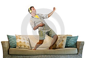 Young happy and excited man jumping on sofa couch listening to music with mobile phone and headphones playing air guitar crazy hav