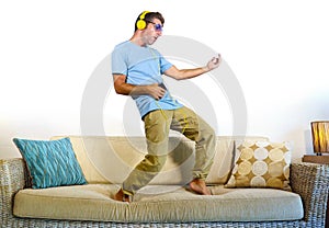 Young happy and excited man jumping on sofa couch listening to music with mobile phone and headphones playing air guitar crazy hav