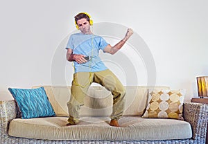 Young happy and excited man jumping on sofa couch listening to music with mobile phone and headphones playing air guitar crazy hav