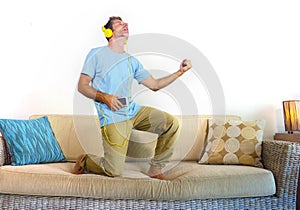 Young happy and excited man jumping on sofa couch listening to music with mobile phone and headphones playing air guitar crazy hav