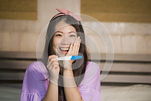 Young happy excited pregnant Asian Chinese woman at home holding predictor and checking positive result on pregnancy test feeling