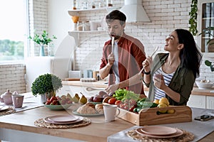 Young happy family couple dancing, singing and having fun while preparing healthy food in the modern kitchen at home