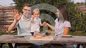 Young Happy Family Having Picnic In The Garden. Mom, Dad And Son Eating Pizza Outdoors