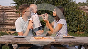 Young Happy Family Having Picnic In The Garden. Mom, Dad And Son Eating Pizza Outdoors