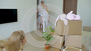 Young happy family moving to new apartment. Two little girls running into new home with parents at background cardboard