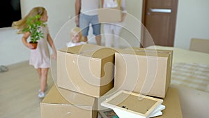 Young happy family moving to new apartment. Two little girls running into new home with parents at background cardboard