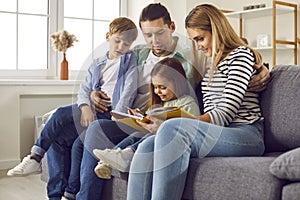 Young happy family reading a book to their two kids sitting on the sofa at home.