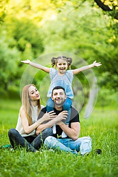 Young happy family of three having fun together outdoor. Pretty little daughter on her father back with happy raised hands. Parent