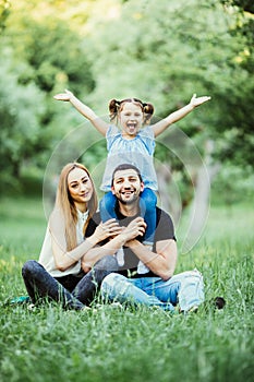 Young happy family of three having fun together outdoor. Pretty little daughter on her father back with happy raised hands. Parent